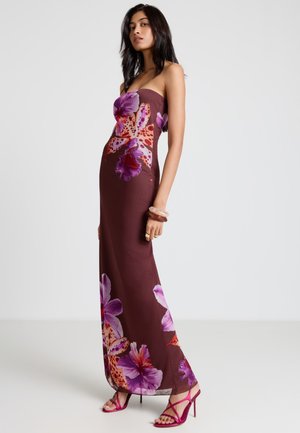 Femme portant une robe longue marron sans bretelles avec de grandes impressions florales violettes et rouges, associée à des sandales roses à talons hauts et des bracelets.