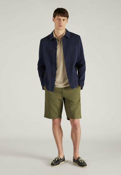 Marineblauw jasje met knoopsluiting, lichtbeige shirt, olijfgroene shorts en zwarte schoenen met witte zolen. Model staat met zijn handen in de zakken.