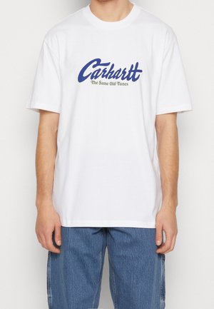 Osoba nosząca białą koszulkę Carhartt z niebieskim tekstem "Carhartt" i mniejszym tekstem "The Same Old Tunes", do tego niebieskie dżinsy.