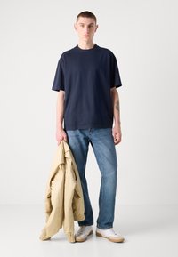 Námořnická bavlněná tričko s kulatým výstřihem, volný střih; nošeno s světle modrými džíny a drženo béžová bunda, doplněno bílými teniskami.