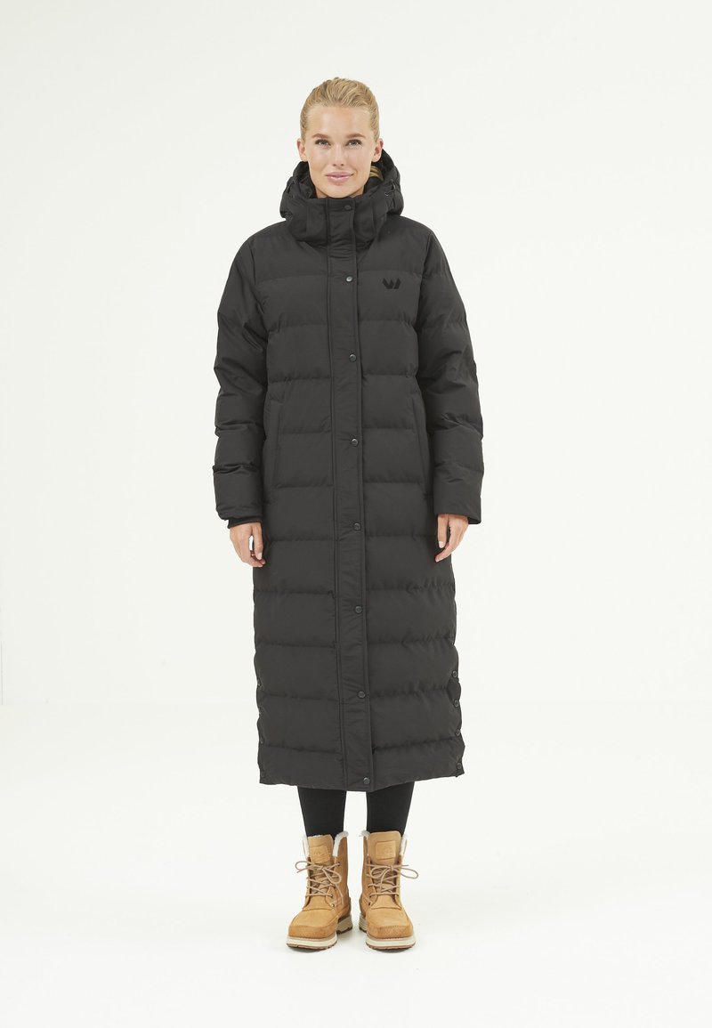 Manteau en doudoune noir avec capuche, de longueur longue, fermeture à boutons sur le devant et texture matelassée. Associé à des bottines beige et des leggings noirs.
