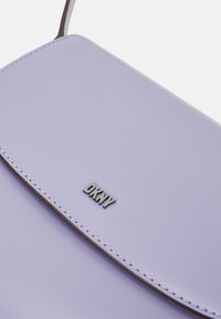 Lila nahkakäsilaukku, jossa on sileä pinta, taitettuna läppänä ja metallinen "DKNY"-logo. Korostettu valkoisella ompelulla.