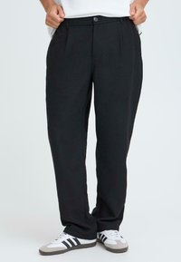 Zwarte broek van lichtgewicht stof met plooien, een knoopsluiting en zijzakken. Draag met witte sneakers met zwarte strepen.