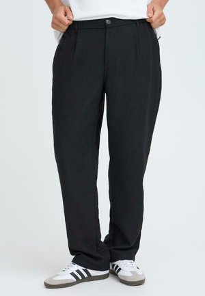 Schwarze Hose aus leichtem Stoff mit Falten, einem Knopfverschluss und Seitentaschen. Kombiniert mit weißen Sneakers mit schwarzen Streifen.