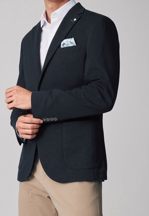 Homme ajustant le poignet d'un blazer bleu marine avec une pochette blanche à motifs, une chemise blanche et un pantalon beige sur un fond gris.
