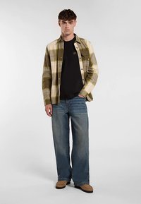 Koszula w zielono-beżową kratę na czarnym t-shircie, w połączeniu z szerokimi niebieskimi jeansami i brązowymi butami. Prosty krój i luźne dopasowanie.