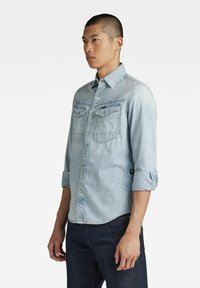 Light blue denim shirt with long sleeves, button-down collar, double chest pockets, and rolled cuffs, paired with dark jeans.
