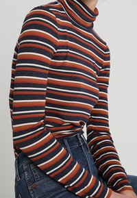 Top de manga larga y cuello alto con rayas horizontales en navy, naranja y crema. Textura acanalada con un diseño ajustado.