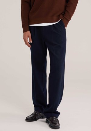 Homme portant un pantalon habillé bleu foncé, des mocassins en cuir noir et un pull marron avec une chemise blanche en dessous, debout les mains dans les poches.