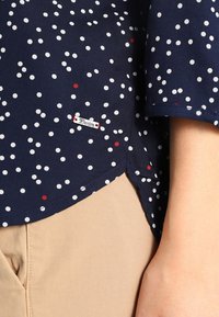 Blusa blu navy con pois bianchi e rossi, maniche corte, orlo curvo e un piccolo etichetta d'argento con scritto "Denim". Pantaloni beige chiaro visibili.