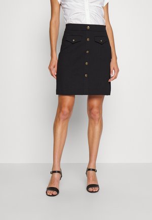 Woman wearing a black buttoned skirt with two front flap pockets and black ankle strap sandals against a plain background.