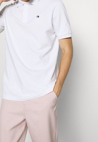 Weißes Baumwoll-Polohemd mit einem geknöpften Kragen und einem kleinen marineblauen Logo auf der linken Brust, kombiniert mit hellrosa Hosen.