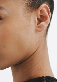 Pendiente de aro plateado en la parte superior de la oreja. Tonalidad de piel suave con un ligero brillo. Primer plano de la oreja y la línea de la mandíbula, mostrando la textura natural.