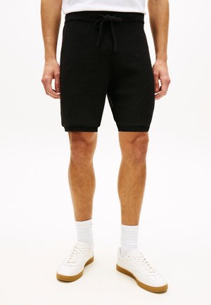 Man draagt zwarte shorts met trekkoord, witte sportsokken en witte sneakers met gumkleurige zolen, staand tegen een effen achtergrond.