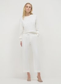 Felpa bianca con sottili decorazioni, abbinata a pantaloni bianchi a gamba dritta. Il modello indossa eleganti scarpe con i piedi scoperti e scintillanti. Design minimalista.