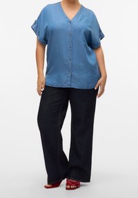 Woman wearing a loose blue button-up short sleeve top, wide black pants, and red open-toe heeled sandals with floral details.