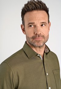 Olivgrünes, strukturiertes Hemd mit Kragen, einer einzelnen Brusttasche und gemusterter Innenauskleidung. Mit weißen Knöpfen und tailliertem Design.