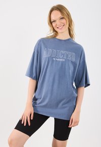 Modra oversized majica z napisom "ODVISNIK OD RAJA" v belem, v kombinaciji z črnimi prilegajočimi kratkimi hlačami. Material se zdi mehak in s sproščenim krojem.