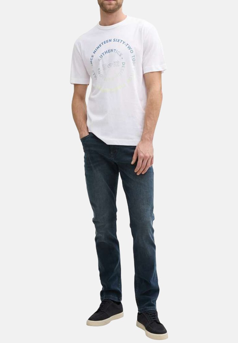 T-shirt en coton blanc à col rond et manches courtes, avec un design de texte circulaire multicolore. Associé à un jean bleu foncé et des baskets noires.