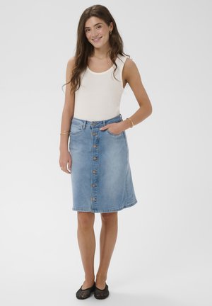 Mladá žena sa usmieva, má na sebe biely tielkový top bez rukávov, riflovú sukňu s gombíkmi po celej dĺžke a čierne baleríny, stojí pred jednoduchým pozadím.