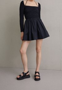 Svart mini-klänning med smockad bodice och långa ärmar, matchad med kraftiga svarta sandaler med flera remmar och spännen.