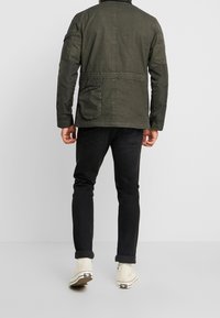 Olivgrön jacka med en dragkedjeficka, texturerat tyg och en avslappnad passform, i kombination med svarta jeans och vita sneakers.