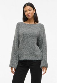 Szary, oversize sweter z teksturowanym, puszystym materiałem, szerokim dekoltem w łódkę i długimi rękawami. W połączeniu z czarnymi dżinsami.