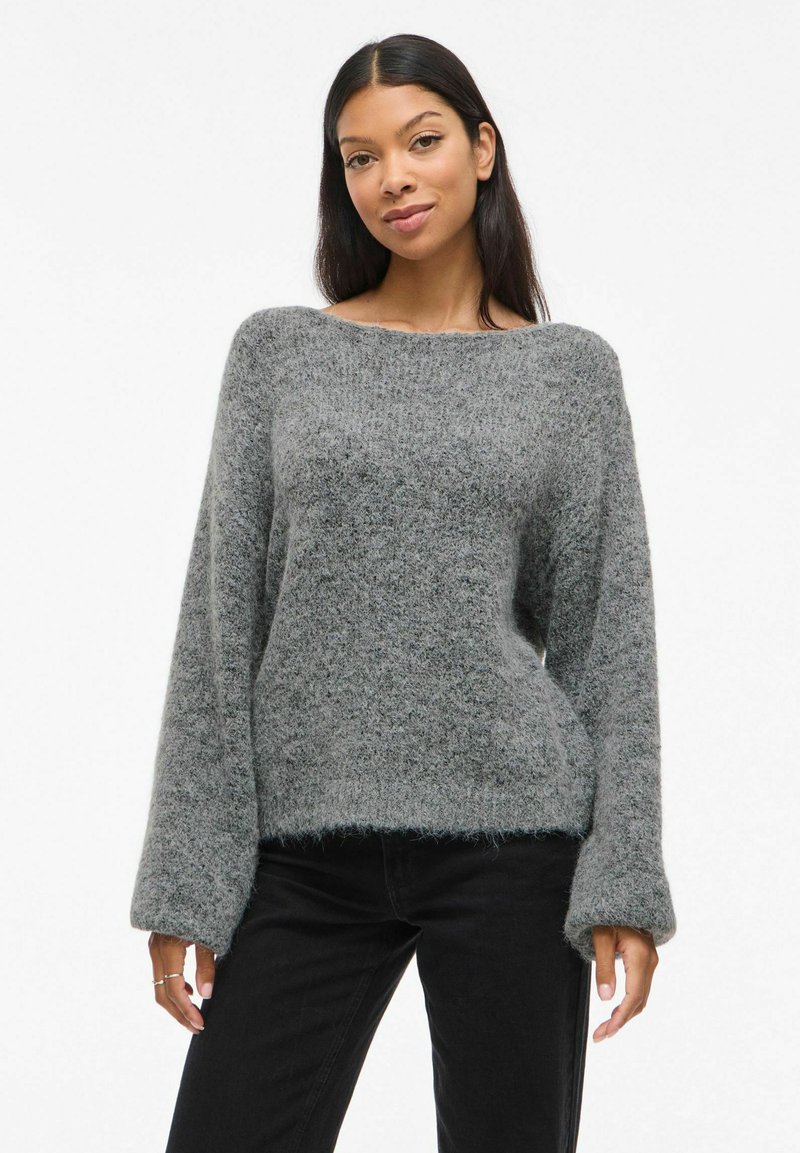Szary, oversize sweter z teksturowanym, puszystym materiałem, szerokim dekoltem w łódkę i długimi rękawami. W połączeniu z czarnymi dżinsami.