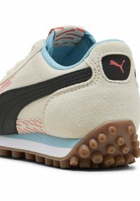 Baskets de sport Puma en crème, avec des bandes en cuir noir, un doublure bleue, des accents roses et une semelle en caoutchouc texturée avec des picots circulaires.