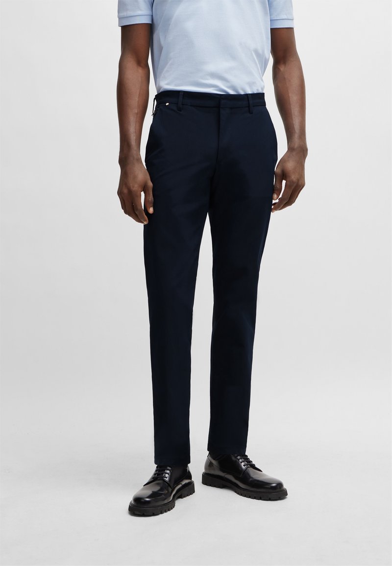 Homme portant un pantalon bleu marine ajusté, une chemise bleu clair à manches courtes et des chaussures de ville noires et cirées, debout sur un fond uni.