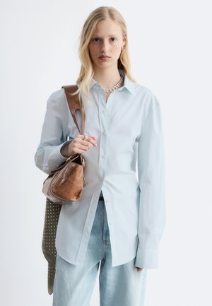 Jeune femme portant une chemise bleu clair à boutons et un jean, tenant un sac à bandoulière en cuir marron avec chaîne sur fond blanc.