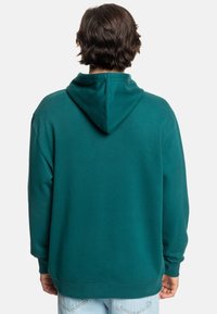 Zelená mikina s kapucňou, vo voľnom strihu, mäkkou textúrou, s rebrovanými manžetami a spodným lemom, pričom má klokanie vrecko a hrubú šnúrku na sťahovanie.