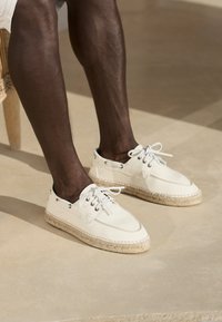 Weiße Espadrille-Schuhe mit einer gewebten Jutesohle, Schnürdesign und metallischen Ösen. Obermaterial aus glattem Leder mit kontrastierenden Steppnähten.