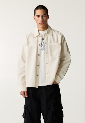 Beige denim jack met een kraag, twee voorzakken en zilveren drukknoopjes, gecombineerd met een witte graphic t-shirt en zwarte cargobroek.