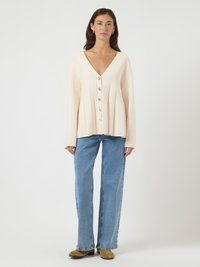 Femeie îmbrăcată cu un cardigan crem cu decolteu în V și butoane, blugi largi albaștri, cercei aurii cerc și bocanci scurți din suede bej, stând în fața unui fundal alb.