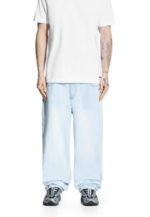 Mężczyzna w luźnych jasnoniebieskich dżinsach, białej koszulce i szaro-czarnych trampkach, stojący z rękami swobodnie opuszczonymi wzdłuż ciała.