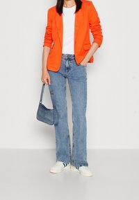 Blazer orange, haut blanc côtelé, jean évasé bleu clair et baskets blanches avec des rayures vertes. Petit sac à main en jean, texture lisse.