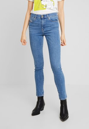 Femme portant un jean skinny bleu clair, des bottines noires et un t-shirt blanc avec un motif jaune et noir, debout devant un fond gris.