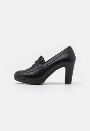 Mocassins à talons en cuir noir avec un bout arrondi, présentant un détail cousu et un talon bloc de hauteur intermédiaire pour la stabilité. Texturé lisse.