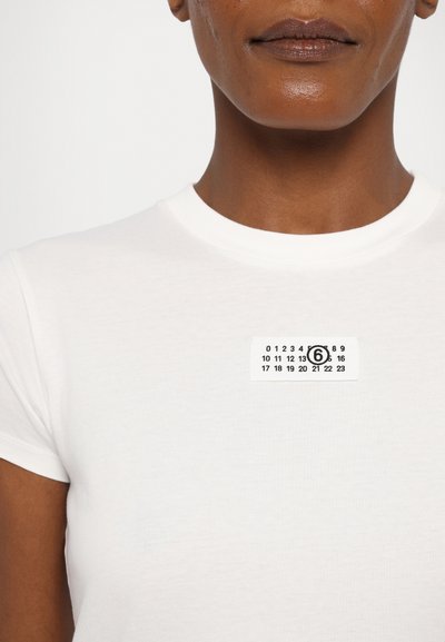 White cotton t-shirt with a round neckline featuring a black and white rectangular patch displaying numbers and the number 6 prominently.