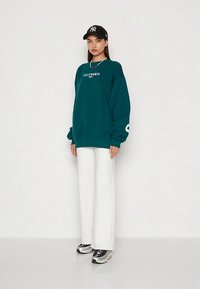 Mujer vestida con un sudadera de gran tamaño de color verde oscuro que dice "California", pantalones anchos blancos, gorra negra y zapatillas, de pie contra una pared blanca.