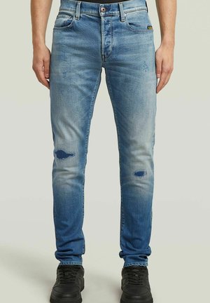 Jasnoniebieskie dopasowane jeansy z wyblakłymi plamami i małymi rozdarciami na kolanach, noszone przez stojącą osobę w czarnych trampkach.