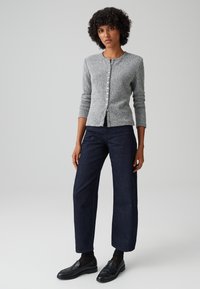 Cardigan grigio lavorato a maglia con maniche lunghe e chiusura a bottoni, abbinato a jeans a gamba larga blu scuro e mocassini neri. Texture liscia e design aderente.