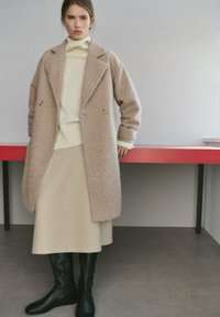 Abrigo de mezcla de lana beige de doble botonadura sobre un suéter crema y una falda a juego, combinado con botas de cuero negro hasta la rodilla. Texturas suaves visibles.