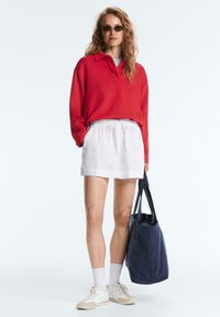 Femeie purtând un tricou roșu cu mâneci lungi, pantaloni scurți albi cu șnur, ochelari de soare, adidași albi, ținând o geantă tip tote albastru închis, stând în fața unui fundal simplu.