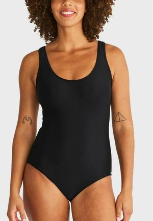 Vrouw met krullend haar die een zwart eendelig badpak draagt, glimlacht en tatoeages van een oog met weegschalen en een driehoek op haar armen toont.