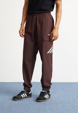 Pantaloni atletici maro dintr-un material neted, cu talie elastică, buzunare laterale și accente albe în dungi. Adidași negri cu trei dungi.