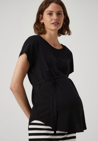 Top maternità nero con maniche corte, scollatura rotonda e vita legata. Il tessuto è liscio con un orlo curvo, abbinato a un fondo a righe.