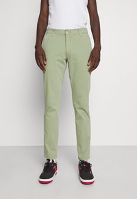 Pantalones de algodón verde claro con un corte ajustado, con cierre frontal de botones y bolsillos laterales, combinados con zapatillas deportivas negras y rosas.