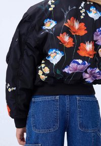 Chaqueta bomber negra con colorido bordado floral en naranja, azul y blanco. Puños y cintura acanalados con un diseño elegante.
